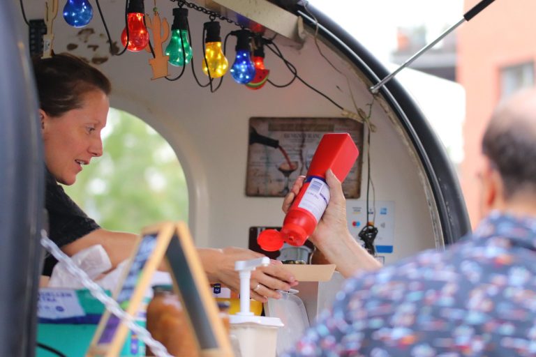 Persoon serveert condimenten vanuit een kleine kraam met kleurrijke verlichting.