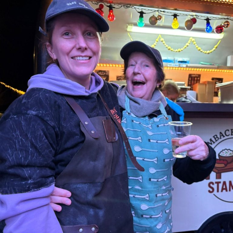 Twee vrouwen in werkoveralls met een drankje, lachend bij een voedselstand.