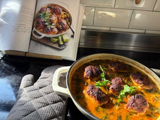 Gekruid vleesgerecht met gehaktballen in een pan, naast een kookboek met een vergelijkbaar gerecht.